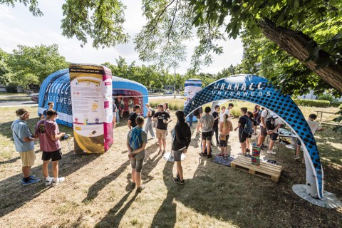zwei neun quadratmeter große Pavillons stehen auf sandigem Boden unter Bäumen. Auf Ihnen steht "Urknall unterwegs" und "Teilchenwelt". Junge leute in sommerlicher Kleidung stehen unter den Pavillons uns betrachten Exponate auf Tischen.