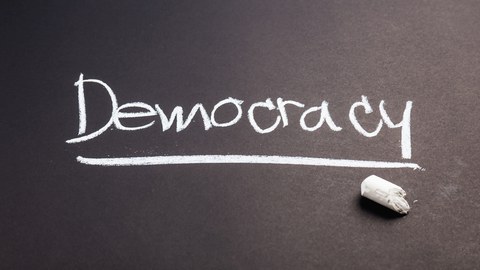 Der Schriftzug "Democracy" wird mit Kreide auf eine Tafel geschrieben. 