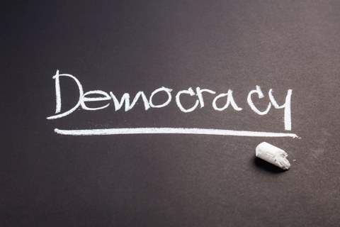 Der Schriftzug "Democracy" wird mit Kreide auf eine Tafel geschrieben. 