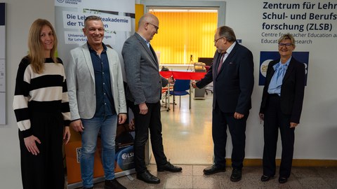 5 Personen stehen in einem Halbkreis dem Fotografen zugewandt. Hinter Ihnen mittig ein Türrahmen und links und rechts zwei Roll-Ups. Außen stehen zwei Frauen der Gruppe, in der Mitte drei Männer. Zwei von ihnen zerschneiden ein Band, dass über die Türzargen gespannt ist.