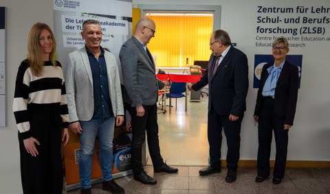 5 Personen stehen in einem Halbkreis dem Fotografen zugewandt. Hinter Ihnen mittig ein Türrahmen und links und rechts zwei Roll-Ups. Außen stehen zwei Frauen der Gruppe, in der Mitte drei Männer. Zwei von ihnen zerschneiden ein Band, dass über die Türzargen gespannt ist.