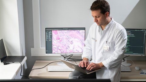 Foto von Prof. Jakob N. Kather, der auf einem Schreibtisch sitzt mit einem Laptop auf dem Schoß. Auf dem Tisch ist ebenfalls ein Computer zu erkennen.