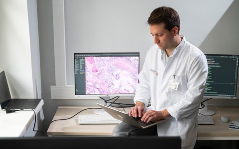 Foto von Prof. Jakob N. Kather, der auf einem Schreibtisch sitzt mit einem Laptop auf dem Schoß. Auf dem Tisch ist ebenfalls ein Computer zu erkennen.