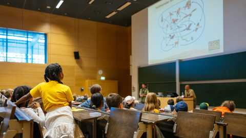 Foto von der Kinder-Universität Dresden: Mehrere Kinder sind in einem Hörsaal der TU Dresden und hören einem Vortrag von zwei Personen zu, die vor einer Tafel an einem Pult stehen.