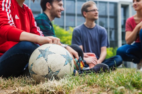 Foto: Vier Studierende sitzen auf einer Wiese am Hörsaalzentrum. Ein Fußball liegt im Vordergrund.