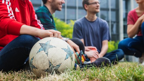 Foto: Vier Studierende sitzen auf einer Wiese am Hörsaalzentrum. Ein Fußball liegt im Vordergrund.