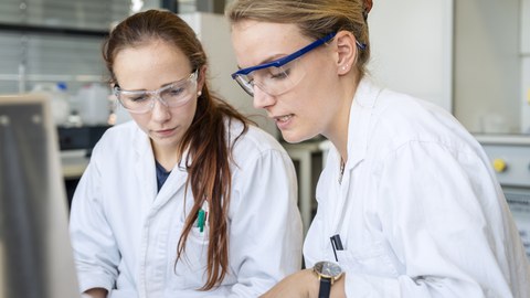 Foto von zwei Frauen im Labor. Beide tragen Arbeitskleidung und Schutzbrillen.