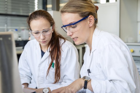 Foto von zwei Frauen im Labor. Beide tragen Arbeitskleidung und Schutzbrillen.