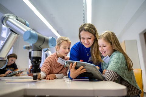 eine Frau lehnt auf einem Tisch und hält ein digitales Tablet in der Hand. Um sie herum stehen zwei junge Mädchen. Alle lächeln und sind auf das Gerät fokussiert. Links neben ihnen steht ein Ribiterarm auf dem Tisch. 