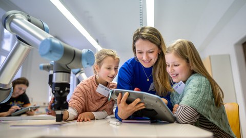 eine Frau lehnt auf einem Tisch und hält ein digitales Tablet in der Hand. Um sie herum stehen zwei junge Mädchen. Alle lächeln und sind auf das Gerät fokussiert. Links neben ihnen steht ein Ribiterarm auf dem Tisch. 