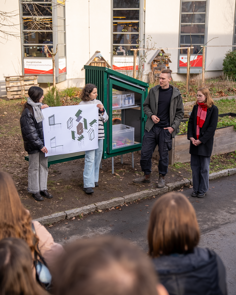 Foto: In einem Garten stehen Menschen vor einer grünen Holzkonstruktion – dem "Fairteiler". Eine Person spricht, andere halten einen Papier in den Händen, das Entwürfe der Konstruktion zeigt.