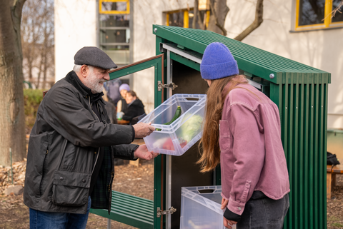 Foto: Zwei Menschen stehen an einer grünen Holzkonstruktion, dessen Tür geöffnet ist. Sie entnehmen eine Kiste, in der Lebensmittel liegen.