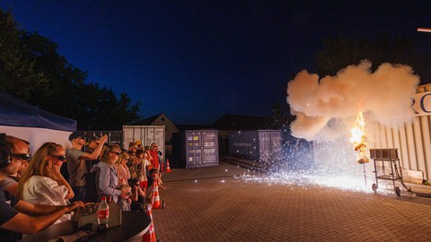Foto einer Puppe, bei der gerade etwas explodiert und in Flammen steht. Aus sicherem Abstand wird die Szene von Zuschauenden beobachtet.