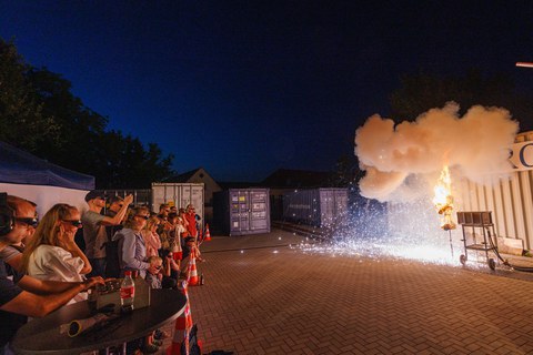 Foto einer Puppe, bei der gerade etwas explodiert und in Flammen steht. Aus sicherem Abstand wird die Szene von Zuschauenden beobachtet.