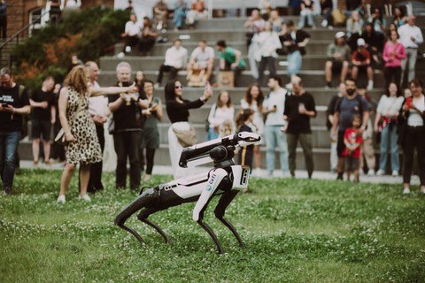 Roboterhund SPOT steht auf der Wiese am Fritz-Foerster-Bau. Im Hintergrund stehen mehrere Menschen in lockerer Atmosphäre ins Gespräch vertieft.