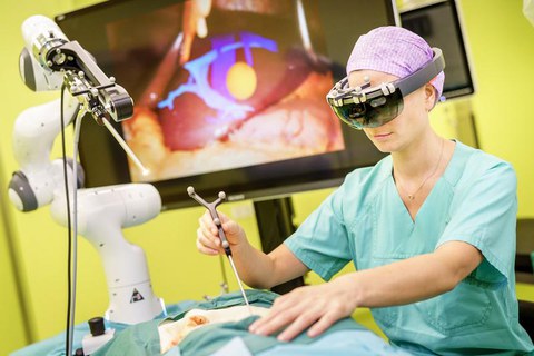 EIne OP-Mitarbeiterin mit Kittel und Häubchen trägt eine VR-Brille und benutzt chirurgisches Werkzeug an einem medizinischen Dummy. Ein Roboterarm unterstützt sie. Auf einem Bildschirm im Hintergrund sieht man das Innere des Dummys im OP-Vorgang, was die Mitarbeiterin in der VR-Brille wahrnimmt.