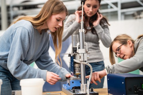 Fotoaufnahme von drei jungen Frauen, die an einer Werkbank stehen, auf dem sich eine Presse befindet.