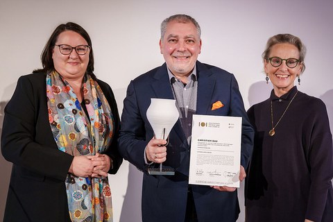 Katrin Stump (links) und Prof.in Ursula Staudinger (rechts) übergeben die Urkunde und Gefäßstatuette an Spitzenkoch Christian Bau.