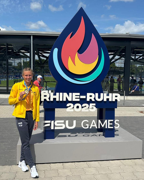Foto von der Leichtathletin Vivienne Morgenstern, die ihre Goldmedaille vor dem Logo der World University Games Rhine-Ruhr 2025 präsentiert.