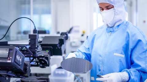 Fotoaufnahme einer Personen in einem Labor für Mikroelektronik. Die Person trägt Kittel, Handschuhe und eine Schutzhaube um den Kopf. Sie hält einen Wafer in der Hand.