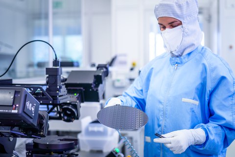 Fotoaufnahme einer Personen in einem Labor für Mikroelektronik. Die Person trägt Kittel, Handschuhe und eine Schutzhaube um den Kopf. Sie hält einen Wafer in der Hand.