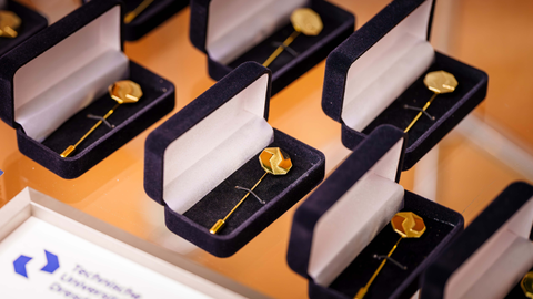 Mehrere geöffnete Schmuckschatullen stehen geordnet auf einer glatten Oberfläche. In jeder Schatulle liegt eine goldfarbene Anstecknadel.