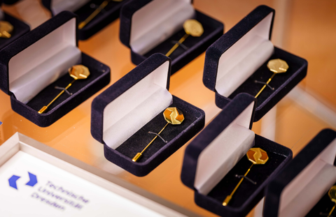 Mehrere geöffnete Schmuckschatullen stehen geordnet auf einer glatten Oberfläche. In jeder Schatulle liegt eine goldfarbene Anstecknadel.