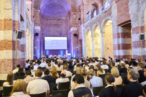 The photograph shows the interior of a church nave, transformed into a large event hall, with an audience and a panel discussion taking place on stage.