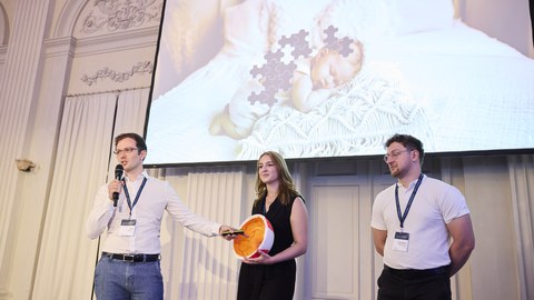 Das Foto zeigt drei Vortragende auf der Bühne. Eine Person spricht ins Mikrofon, während ein Augenmodell präsentiert wird.