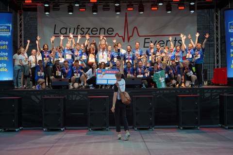 Eine große Gruppe von Teilnehmenden eines Laufevents steht auf einer Bühne und jubelt mit erhobenen Armen. Viele tragen Medaillen und farbige Laufshirts. Vor der Bühne steht eine Person mit Kamera und fotografiert die Gruppe.