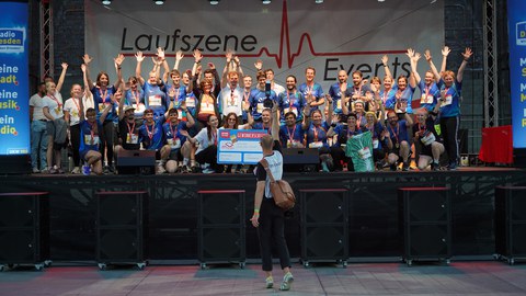 Eine große Gruppe von Teilnehmenden eines Laufevents steht auf einer Bühne und jubelt mit erhobenen Armen. Viele tragen Medaillen und farbige Laufshirts. Vor der Bühne steht eine Person mit Kamera und fotografiert die Gruppe.