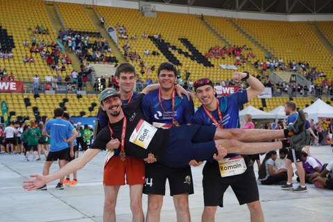 Vier Personen posieren lachend im Innenraum eines Stadions. Drei von ihnen heben eine weitere Person waagerecht in die Luft. Alle tragen Sportkleidung, Startnummern und Medaillen. Im Hintergrund sitzen und stehen zahlreiche weitere Teilnehmende und Zuschauende auf den gelben Tribünenrängen.