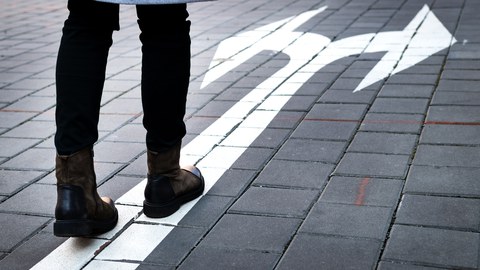 Unterkörper einer Person, die auf einem gepflasterten Weg steht. Vor den Schuhen ist ein weißer Pfeil auf den Boden gemalt, der sich nach vorne gabelt und zwei Richtungen anzeigt.