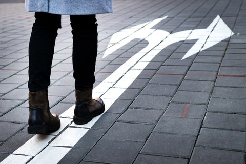 Unterkörper einer Person, die auf einem gepflasterten Weg steht. Vor den Schuhen ist ein weißer Pfeil auf den Boden gemalt, der sich nach vorne gabelt und zwei Richtungen anzeigt.