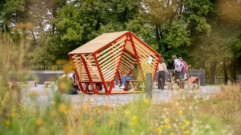 Foto: Eine offene, geometrische Holzstruktur mit roten Rahmen und schräg angeordneten hellen Latten steht auf einer Kiesfläche inmitten von Wiesenpflanzen. Eine Person sitzt entspannt im Inneren der Konstruktion, andere stehen seitlich daneben und unterhalten sich. Im Hintergrund befindet sich dichter grüner Baumbestand.