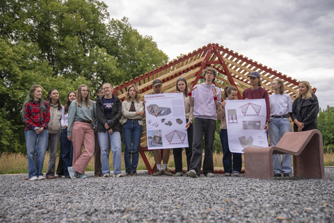 Foto: Eine größere Gruppe junger Menschen steht im Halbkreis vor einer offenen, rot‑gelben Holzstruktur im Freien. Zwei Personen halten große Poster mit architektonischen Entwürfen und erklären etwas, während die übrigen zuhören. Der Hintergrund zeigt Bäume und einen bewölkten Himmel, der Boden besteht aus feinem Kies.