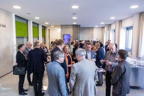 Eine große Gruppe von Menschen steht in einem hellen Foyer bei einer Veranstaltung. Viele unterhalten sich in kleinen Gruppen, halten Getränke oder Teller in der Hand und wirken in Gespräche vertieft. Im Hintergrund sind hohe Fenster, grüne Wandpaneele und ein Buffetbereich zu sehen.