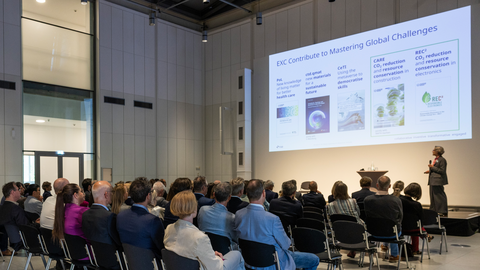 Ein Publikum sitzt in Reihen in einem modernen Vortragssaal und hört TUD-Rektorin Prof. Ursula M. Staudinger zu, die vorn auf der Bühne steht und eine Präsentation zeigt. Auf der großen Leinwand sind mehrere Folienelemente zu sehen, die sich mit Forschung und globalen Herausforderungen befassen.
