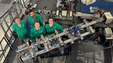 Blick von oben in eine Halle mit experimentellen Aufbauten, davor 4 Personen in grünen Arbeitskitteln