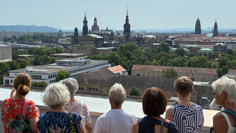 Fotoaufnahme von mehreren älteren Personen auf der Dachterrasse des Hauses der Presse in Dresden. Die Personen blicken auf die Silhoutte der Dresdner Altstadt.
