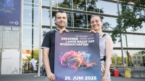 Foto von einem Mann und einer Frau, die vor dem Hörsaalzentrum der TUD stehen und ein Veranstaltungsplakat der Langen Nacht der Wissenschaften 2026 halten.