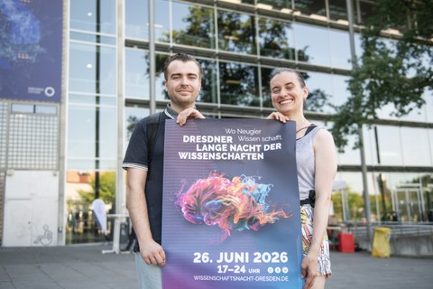 Foto von einem Mann und einer Frau, die vor dem Hörsaalzentrum der TUD stehen und ein Veranstaltungsplakat der Langen Nacht der Wissenschaften 2026 halten.