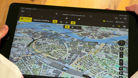 Fotoaufnahme von einem Tablet, auf dem der digitale Zwilling der Landeshauptstadt Dresden zu sehen ist. Der Bildausschnitt zeigt ein 3D-Modell der Innenstadt von Dresden.