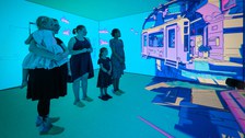 Mehrere Erwachsene und Kinder stehen in einem immersiven Raum mit vollständig blauen Wänden, auf die eine lebhafte, gezeichnete Stadtlandschaft projiziert wird. Alle betrachten die animierte Szene aufmerksam, während sie auf Filzpantoffeln auf dem Boden stehen.