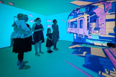 Mehrere Erwachsene und Kinder stehen in einem immersiven Raum mit vollständig blauen Wänden, auf die eine lebhafte, gezeichnete Stadtlandschaft projiziert wird. Alle betrachten die animierte Szene aufmerksam, während sie auf Filzpantoffeln auf dem Boden stehen.