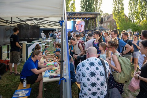 Viele Menschen besuchen einen Outdoor‑Wissenschaftsstand. Unter einem Pavillon sitzen mehrere Kinder und Erwachsene an Tischen und bauen mit bunten Bausteinen, während daneben eine Person an einem Monitor steht. Rechts drängen sich zahlreiche Besuchende vor der Station, im Hintergrund ist eine Bühne mit einer großen LED‑Leinwand zu sehen.