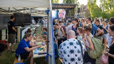 Viele Menschen besuchen einen Outdoor‑Wissenschaftsstand. Unter einem Pavillon sitzen mehrere Kinder und Erwachsene an Tischen und bauen mit bunten Bausteinen, während daneben eine Person an einem Monitor steht. Rechts drängen sich zahlreiche Besuchende vor der Station, im Hintergrund ist eine Bühne mit einer großen LED‑Leinwand zu sehen.