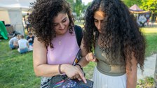 Zwei Personen stehen im Gras und betrachten gemeinsam ein bunt gestaltetes Programmheft. Beide beugen sich über die Seite und zeigen auf einen Punkt im Heft, während sie lächelnd darüber sprechen. Hinter ihnen sitzen weitere Menschen im Schatten.