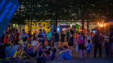 Bei einsetzender Dämmerung sitzen und stehen viele Menschen auf einer Wiese zwischen Bäumen und beleuchteten Ständen. Einige entspannen in Liegestühlen, andere unterhalten sich oder laufen durch die Menge. Im Hintergrund sind bunte Wandmalereien und Lichterdekorationen zu sehen, was der Szene eine sommerliche, festivalartige Atmosphäre verleiht.