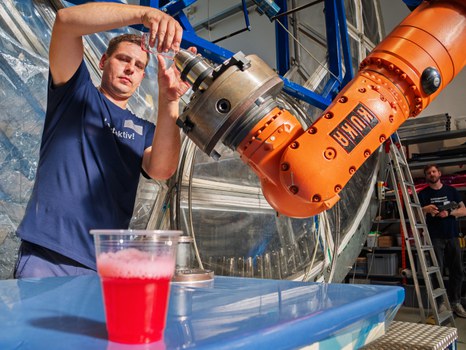 In einer großen Werkhalle demonstriert eine Person die Arbeit an einem orangefarbenen Industrieroboter, indem sie ein Bauteil einspannt. Im Vordergrund steht ein Plastikbecher mit rotem Getränk. Eine weitere Person beobachtet die Szene aus einiger Entfernung.